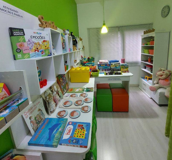Sala infantil com prateleiras de livros e jogos sobre emoções, pufes coloridos e mesa de atividades